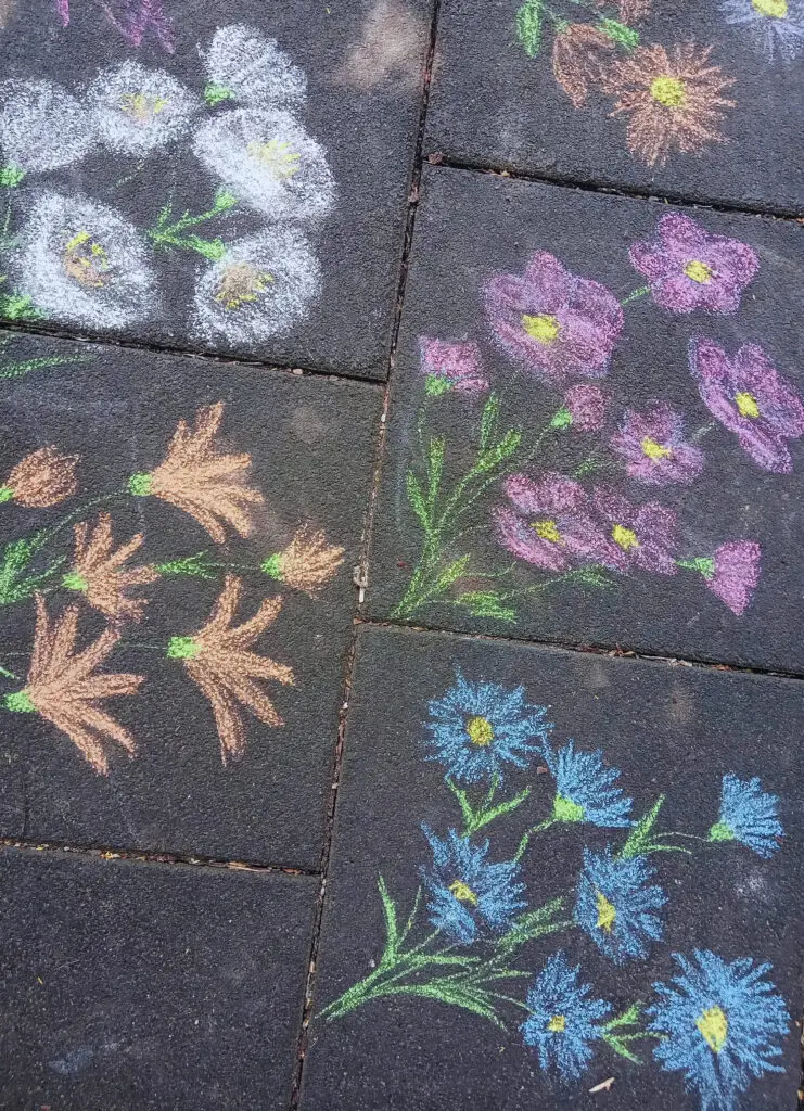 Faint chalk flowers etched by playful hands, fading in the sun and peeking through pavement cracks like echoes of a forgotten afternoon.